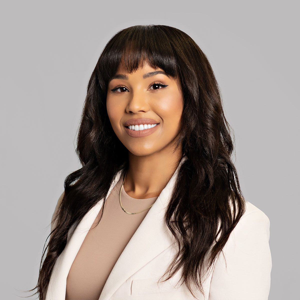 A person with long dark brown hair smiles a the camera. They are wearing a white blazer with a beige blouse underneath and a silver necklace.