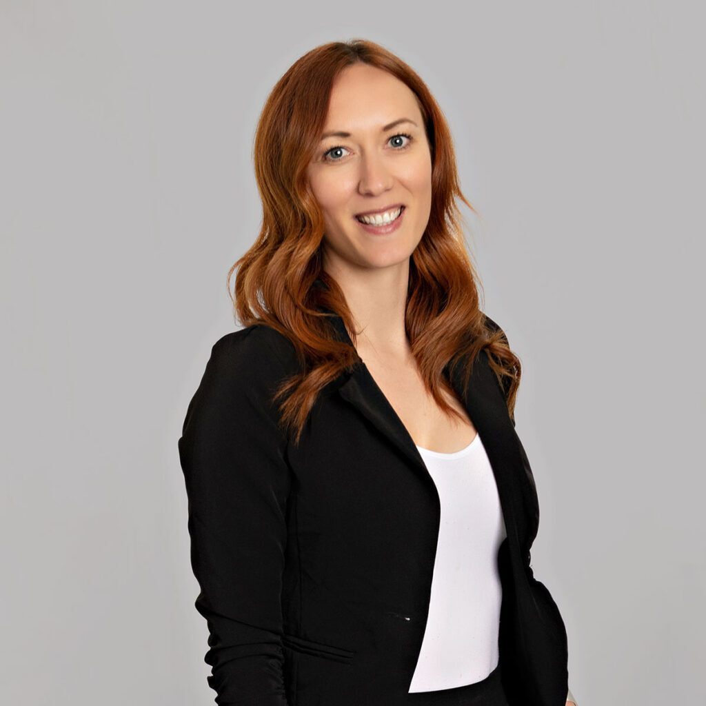Tellia A person with long red hair smiles at the camera. They are wearing a black blazer with a white blouse underneath.